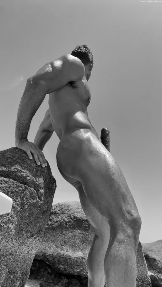 Nude muscular man standing on rocks in sunlight, body wet, viewed from low angle with erection visible